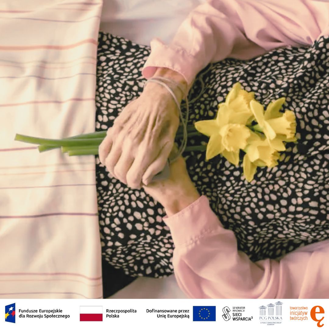 Na zdjęciu znajduje się leżąca zmarła osoba w&nbsp;trakcie pogrzebu domowego. Widać jej tułów i&nbsp;ręce splecione na brzuchu. Jest ubrana w&nbsp;czarną bluzkę w&nbsp;białe kropki i&nbsp;różowy sweterek. Od pasa w&nbsp;dół jest przykryta pościelą. W&nbsp;splecionych dłoniach trzyma bukiet żółtych żonkili.